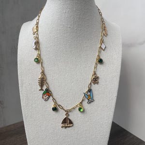 May include: A gold chain necklace with multiple charms, including a sailboat, a fish bone, a cartoon character, and green gemstones.