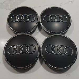 May include: Four black and silver Audi wheel center caps. Each cap features the Audi logo, a set of four interlocking rings, in silver against a black background. The caps have a shiny silver trim.
