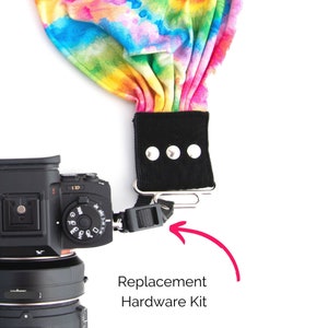 May include: A close-up of a camera strap with a colorful fabric design. The strap has a black adjustable buckle and a metal clip. The text "Replacement Hardware Kit" is written below the clip.