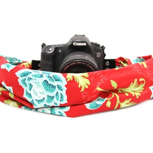 May include: A red camera strap with a floral pattern featuring turquoise and yellow flowers. A black Canon camera is sitting on top of the strap.