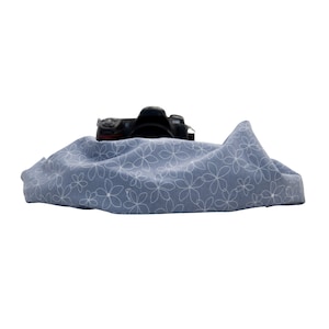 May include: A black DSLR camera rests on a light blue cloth with a white floral pattern. The cloth is draped, creating soft folds. The camera's lens is visible, and the overall composition is clean and simple.