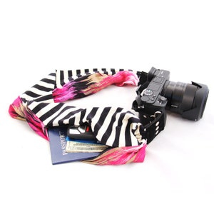 May include: A black and white striped camera strap with a pink and white floral design. The strap is wrapped around a black camera with a lens. A passport, credit cards, and cash are tucked into the strap.