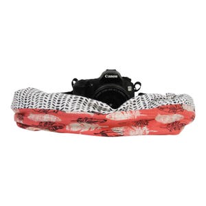 May include: A black Canon DSLR camera rests inside a patterned camera bag. The bag features a coral-colored fabric with feather designs and a black and white dotted pattern. The camera strap is visible.