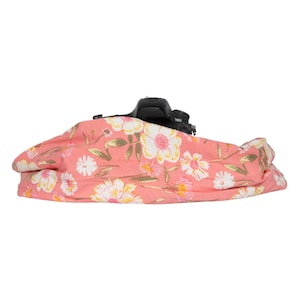 May include: A black DSLR camera rests on a coral-colored floral patterned camera wrap. The wrap features white and yellow flowers with green stems and leaves. The camera is partially visible.