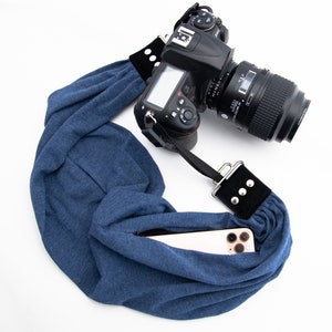 May include: A blue camera strap with a black accent and silver rivets is attached to a black DSLR camera. A smartphone is in the strap's pocket. The strap is made of soft, flexible material.