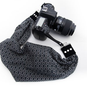 May include: A black DSLR camera with a patterned camera strap. The strap is black with a white geometric design. The strap has black suede-like pads with three white dots on each end. The camera lens is black.