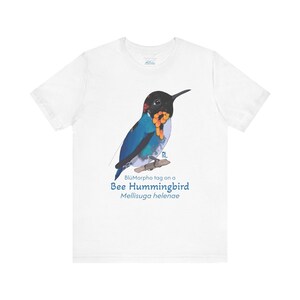 May include: White t-shirt featuring a detailed illustration of a Bee Hummingbird with blue, black, orange, and red plumage. The text "BlüMorpho tag on a Bee Hummingbird Mellisuga helenae" is printed below the bird.