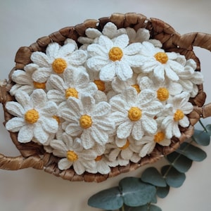 Op de afbeelding: Een rieten mand gevuld met gehaakte witte madeliefjes met gele centra. De mand is bruin en heeft een geweven textuur.