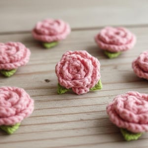 Puede incluir: Primer plano de varias rosas de ganchillo hechas a mano en un tono rosa suave. Cada rosa tiene una base de hoja verde, sobre una superficie de madera clara. Ideal para manualidades y adornos.