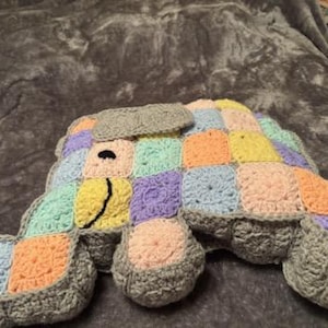 May include: A crocheted elephant pillow with a patchwork design in pastel colors, including pink, yellow, green, and purple. The elephant has a gray body and a black embroidered smile.