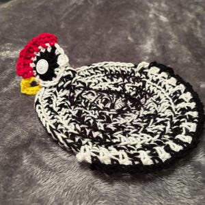 May include: A crocheted black and white chicken dishcloth with a red, white, and yellow crocheted head. The chicken has a black button for an eye.