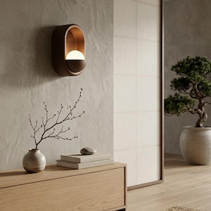 May include: A wall-mounted sconce featuring an oval-shaped dark brown frame with a recessed interior containing a glowing white spherical light bulb and a series of vertical arched lines, providing warm illumination against a textured beige wall.