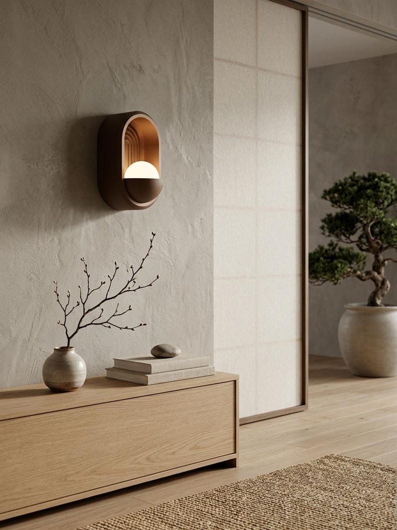 May include: A wall-mounted sconce featuring an oval-shaped dark brown frame with a recessed interior containing a glowing white spherical light bulb and a series of vertical arched lines, providing warm illumination against a textured beige wall.