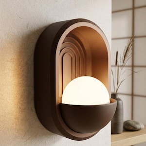 May include: A wall-mounted sconce featuring a matte brown oval frame with an arched interior design. A glowing white spherical light bulb sits within a semi-circular brown bowl at the base of the fixture, casting a warm light against the wall.
