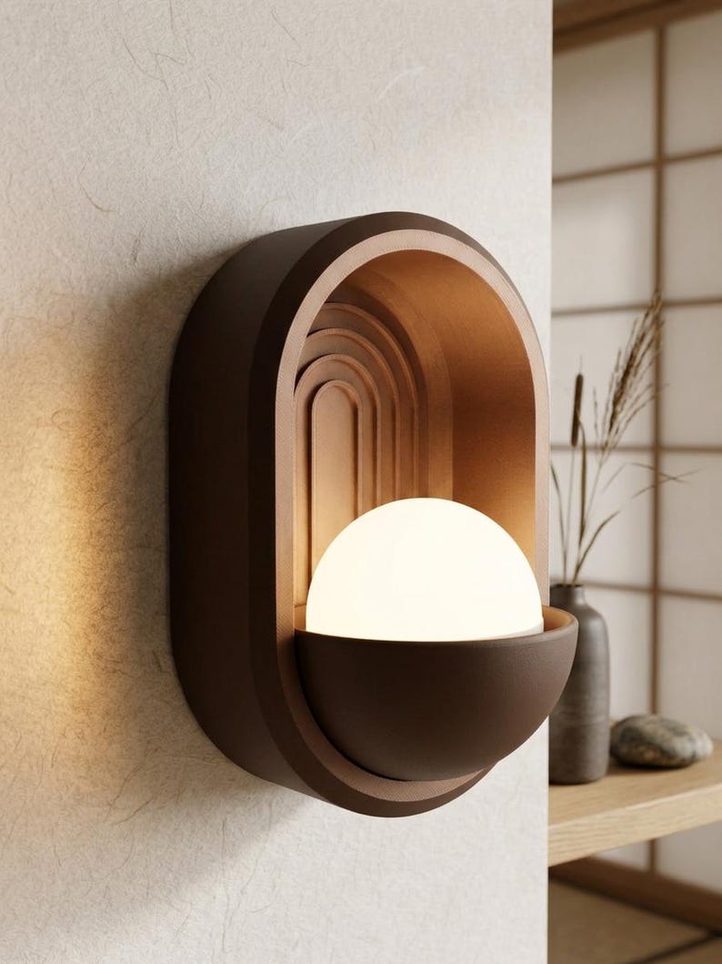 May include: A wall-mounted sconce featuring a matte brown oval frame with an arched interior design. A glowing white spherical light bulb sits within a semi-circular brown bowl at the base of the fixture, casting a warm light against the wall.