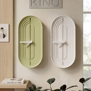 May include: Two oval wall clocks, one in sage green and one in white, featuring concentric grooved lines and white clock hands. Above the clocks, a rectangular sign displays the word KINO in embossed capital letters on a light background.
