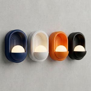 May include: Four wall-mounted sconces arranged in a row, each featuring an oval frame with a recessed arched design and a protruding semi-spherical base. The fixtures are colored navy blue, white, orange, and black, each holding a glowing white orb.
