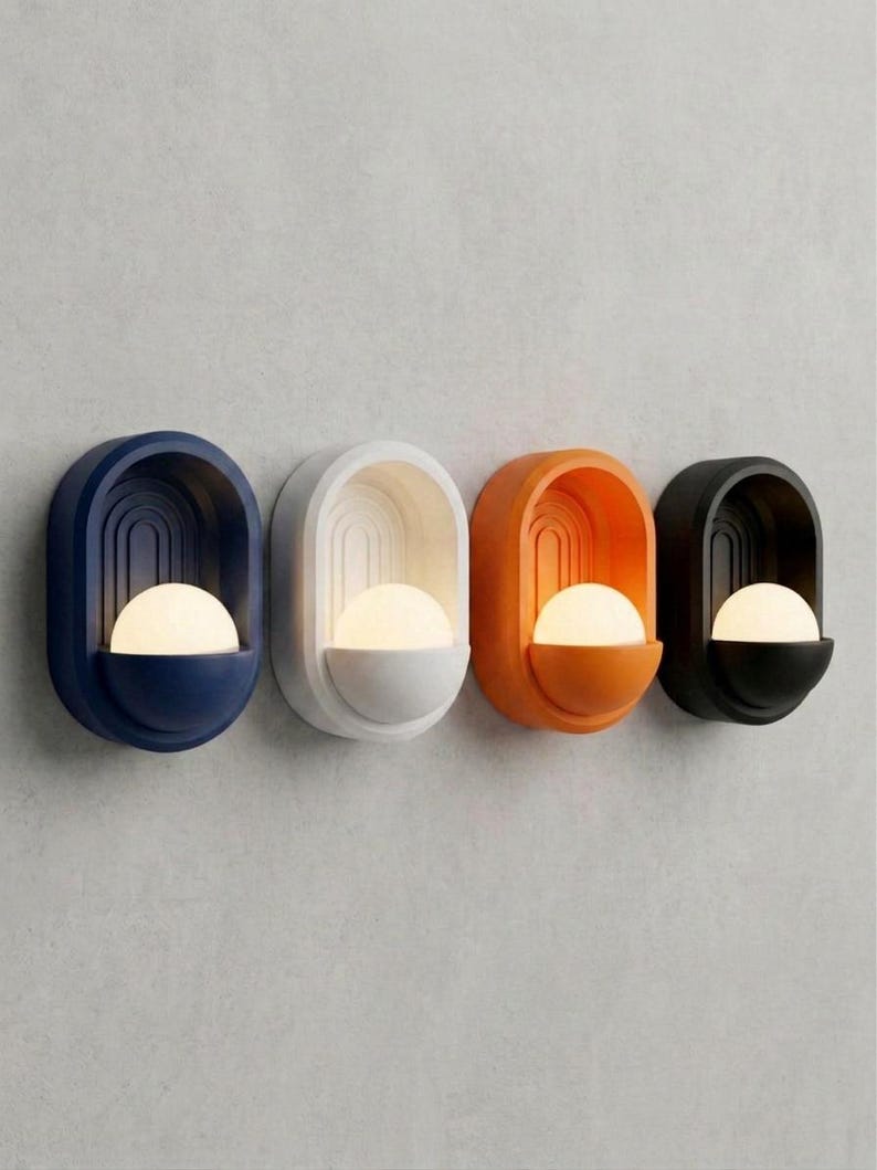 May include: Four wall-mounted sconces arranged in a row, each featuring an oval frame with a recessed arched design and a protruding semi-spherical base. The fixtures are colored navy blue, white, orange, and black, each holding a glowing white orb.