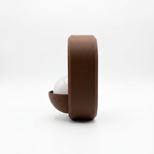 May include: A brown, rounded, rectangular wall lamp featuring a matte finish. A semi-circular brown shade protrudes from the side, housing a white, spherical light bulb that is partially visible within the structure of the fixture.