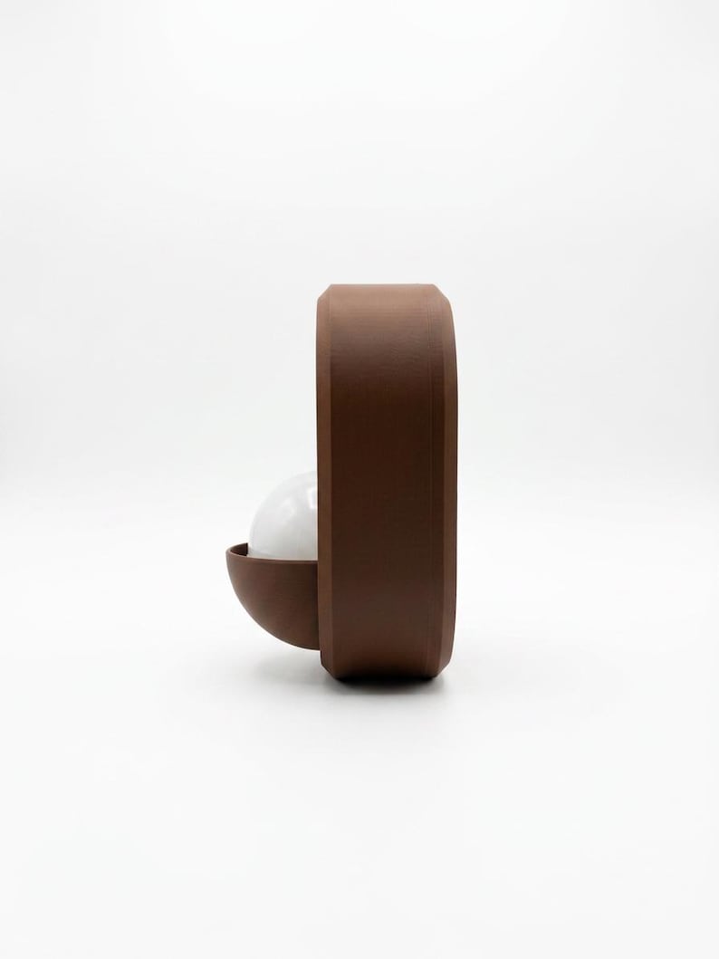 May include: A brown, rounded, rectangular wall lamp featuring a matte finish. A semi-circular brown shade protrudes from the side, housing a white, spherical light bulb that is partially visible within the structure of the fixture.
