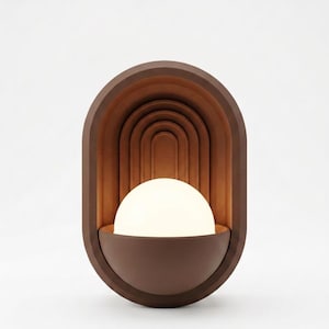 May include: A wall-mounted lamp featuring a dark brown oval frame with a series of concentric arched indentations. A semi-spherical white glass globe sits in the lower portion of the frame, emitting a soft, warm glow that illuminates the interior.