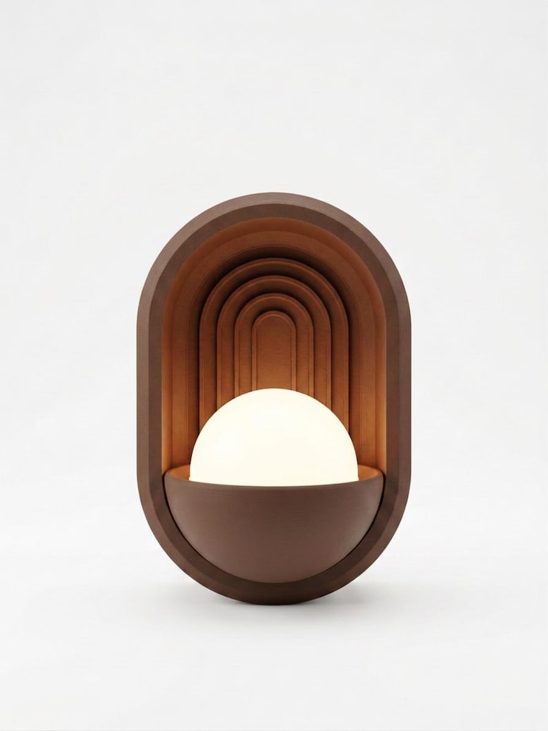 May include: A wall-mounted lamp featuring a dark brown oval frame with a series of concentric arched indentations. A semi-spherical white glass globe sits in the lower portion of the frame, emitting a soft, warm glow that illuminates the interior.