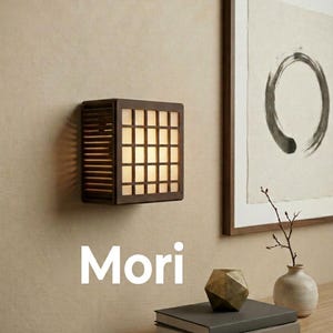 May include: A square wall-mounted lamp with a dark brown wooden frame and a grid-patterned translucent shade that emits a warm glow. The word Mori is printed in white text below the lamp. The lamp features horizontal slats on the side.