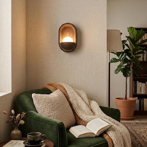 May include: An oval wall sconce with a dark brown frame and concentric arched interior detailing, featuring a central glowing spherical light bulb. The fixture is mounted on a textured beige wall above a green velvet armchair with a knit throw.