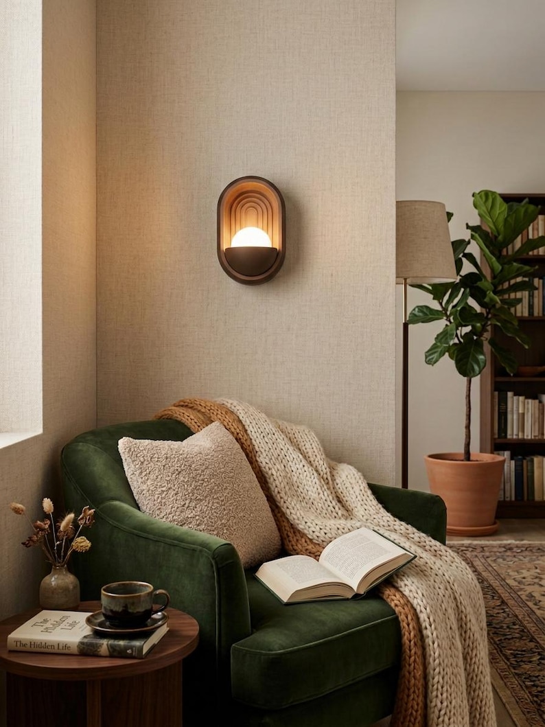 May include: An oval wall sconce with a dark brown frame and concentric arched interior detailing, featuring a central glowing spherical light bulb. The fixture is mounted on a textured beige wall above a green velvet armchair with a knit throw.