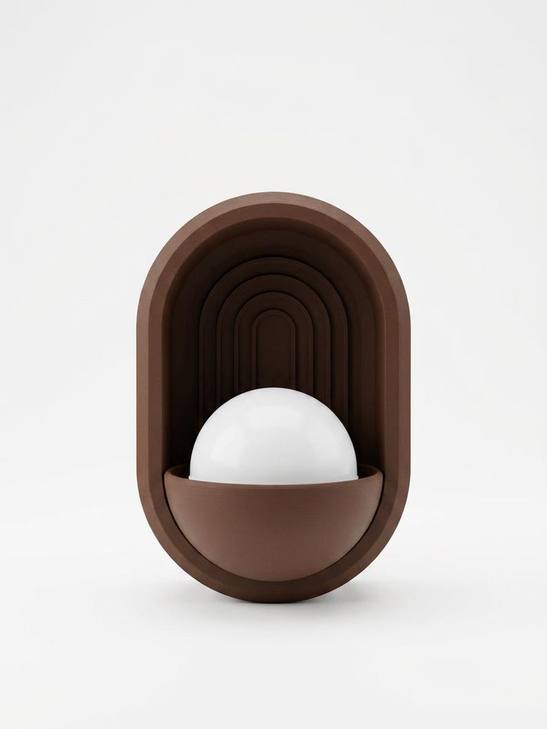 May include: A wall-mounted lamp featuring a matte brown oval frame with a recessed arched interior design. A white hemispherical glass globe sits within a matching brown semi-circular base at the bottom of the fixture, creating a layered look.