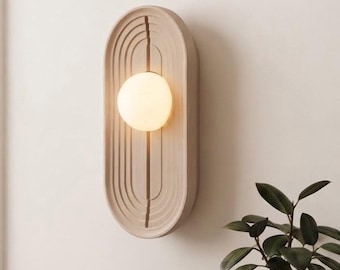 May include: An oval wall sconce with a beige, grooved, concentric arch design and a central vertical slit. A spherical, frosted white glass light bulb is mounted in the center, emitting a warm glow against the light-colored wall surface.