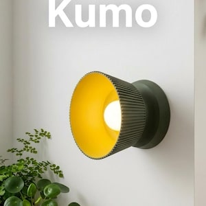 May include: A contemporary wall light with a dark green, ribbed cylindrical shade and a bright yellow interior. The word "Kumo" is displayed in white text above the light. The light is mounted on a white wall, with greenery in the lower left corner.