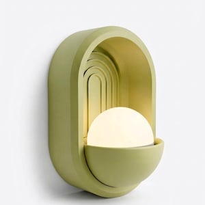 May include: A wall-mounted light fixture featuring a matte sage green arched frame with recessed concentric semi-circular detailing. A glowing white spherical glass bulb sits within a matching semi-circular basin at the base of the structure.