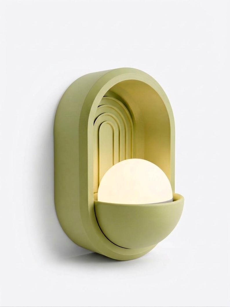 May include: A wall-mounted light fixture featuring a matte sage green arched frame with recessed concentric semi-circular detailing. A glowing white spherical glass bulb sits within a matching semi-circular basin at the base of the structure.