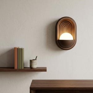May include: A wall-mounted sconce featuring a dark brown oval frame with concentric arched detailing. A semi-circular base holds a glowing white spherical light bulb, creating a warm illumination within the recessed structure of the fixture.