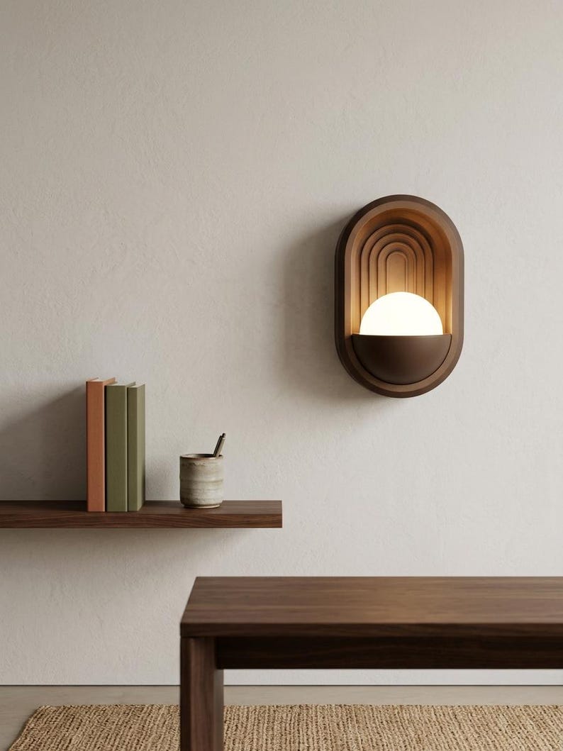 May include: A wall-mounted sconce featuring a dark brown oval frame with concentric arched detailing. A semi-circular base holds a glowing white spherical light bulb, creating a warm illumination within the recessed structure of the fixture.
