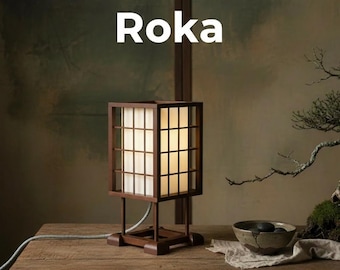 May include: A square wooden table lamp with a grid-patterned frame and a white translucent shade. The word Roka is printed in white text above the lamp. A light gray power cord extends from the base of the lamp, which sits on a wooden surface.