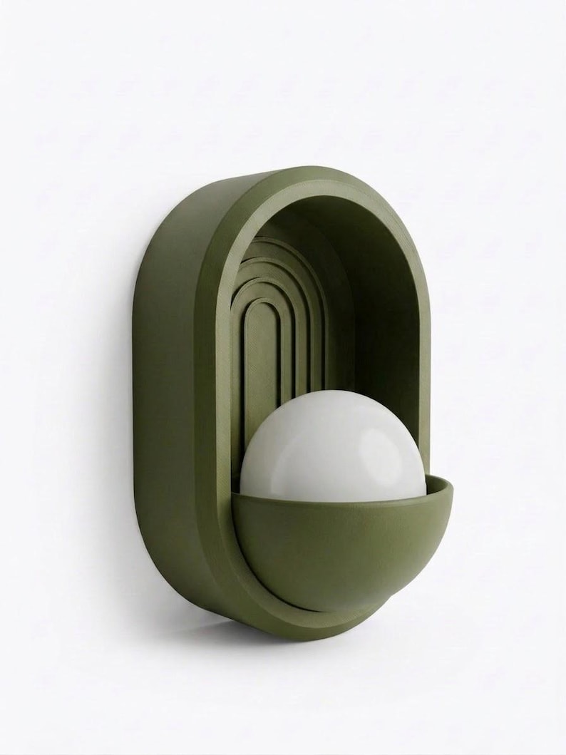 May include: An olive green wall sconce featuring an arched, recessed back panel with concentric semi-circular grooves. A semi-spherical, matte olive green base holds a smooth, opaque white spherical light bulb in the center of the fixture.