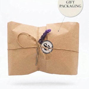 May include: A rectangular package wrapped in brown kraft paper, tied with thin twine. A sprig of purple lavender is tucked under the twine next to a circular white sticker featuring a black ampersand symbol. A circular label above reads GIFT PACKAGING.