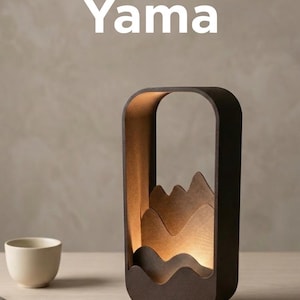 May include: A dark brown, rectangular lamp with a mountain silhouette design. The word "Yama" is printed in white at the top. A small white cup, a notebook, and a pen are on the table.