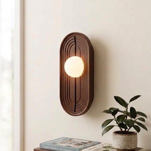 May include: A wall-mounted oval light fitting featuring a dark brown wooden frame with concentric grooved lines and a central spherical white glass bulb. Below, a wooden shelf holds books titled Quantum Mechanics: An Introduction, Molecular Gastronomy Handbook, and a card labelled Galactic Collectibles, alongside a small potted plant.