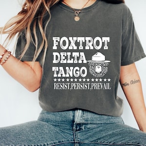 Può includere: Una t-shirt color antracite con un design grafico bianco. La grafica presenta il testo "FOXTROT DELTA TANGO" con un'illustrazione stilizzata di un cappello da ranger e un orso. Il testo "RESIST, PERSIST, PREVAIL" si trova sotto la grafica.