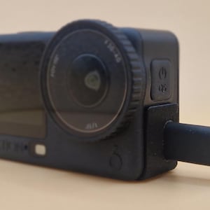 Puede incluir: Una cámara de acción negra con una lente prominente y un botón de encendido. La cámara está conectada a un cable de carga negro. La lente tiene las marcas 4.0-4.6.