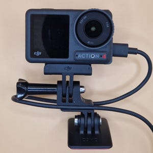 May include: Black DJI Action 4 camera with a lens, screen, and attached mount. The camera is connected to a black mount and base with a black cable. The camera is mounted on a beige surface.