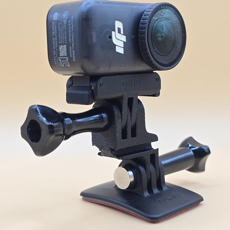 May include: A black DJI Osmo action camera mounted on a black plastic adjustable tripod adapter with a flat adhesive base. The camera features a circular lens, a side-mounted logo, and regulatory text printed on the side of the rectangular housing.