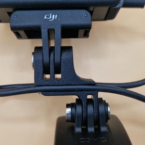 May include: Close-up of a black camera mount with a black cable attached. The mount has a rectangular base with the DJI logo and a series of slots and holes for attachment. The word "OSMO" is visible on the bottom.