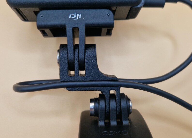 May include: Close-up of a black camera mount with a black cable attached. The mount has a rectangular base with the DJI logo and a series of slots and holes for attachment. The word "OSMO" is visible on the bottom.