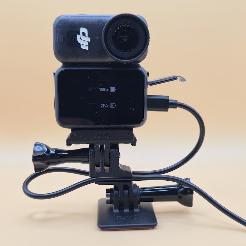 May include: A black DJI Osmo action camera mounted on a base with a charging cable connected to the side. The screen displays battery levels of 100% and 21% with icons. The device features a prominent circular lens and the DJI logo on the front.