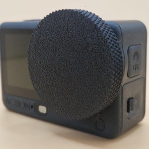 Op de afbeelding: Een zwarte actiecamera met een getextureerde lensdop. De camera heeft een scherm aan de achterkant en knoppen aan de zijkant. Het woord "ACTION" staat op de voorkant.