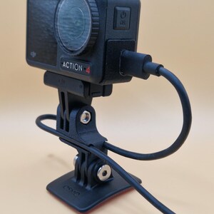 May include: Black action camera labeled "ACTION-4" with a lens and power button, mounted on a black bracket with a cable attached. The camera is connected to a black base with a red adhesive.